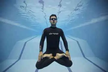 Freediver wearing black wetsuit with legs crossed and eyes closed meditating underwater.