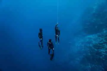 Three freedivers wearing all black freediving gear floating in circle facing inward underwater in blue ocean.
