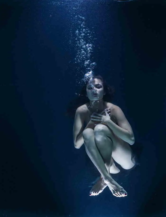 Naked woman sinking underwater clutching legs up to chest and blowing air bubbles.